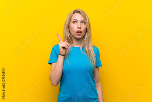 Young blonde woman over isolated yellow background thinking an idea pointing the finger up
