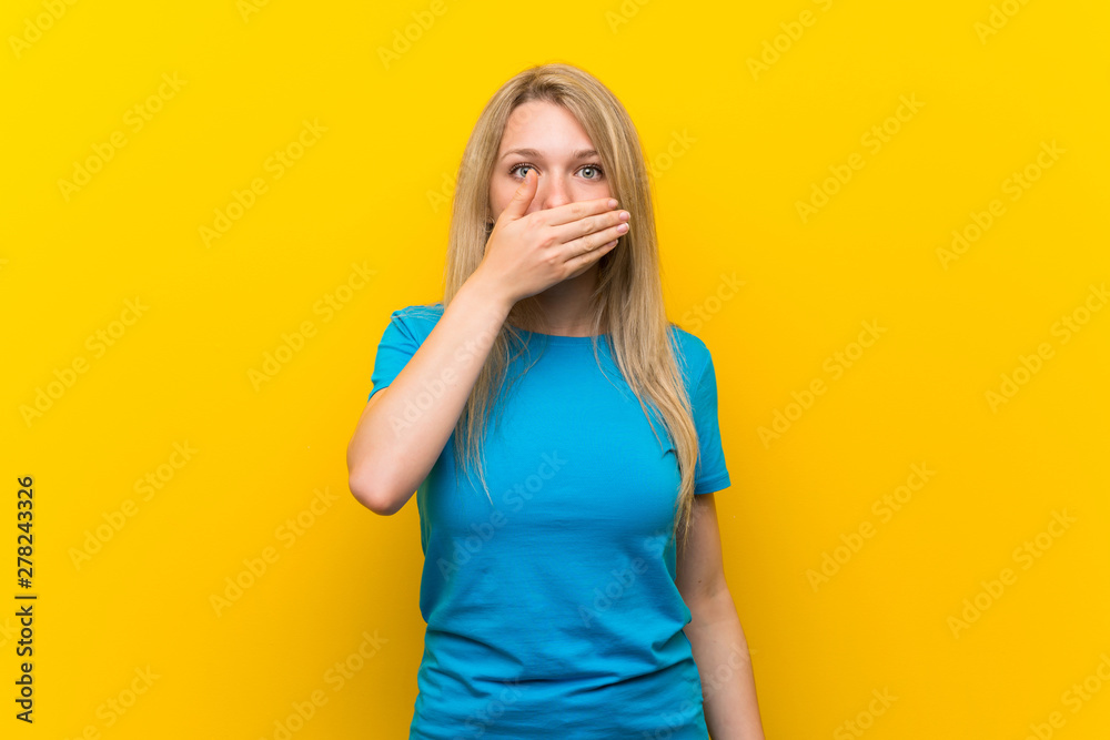 Young blonde woman over isolated yellow background covering mouth with hands