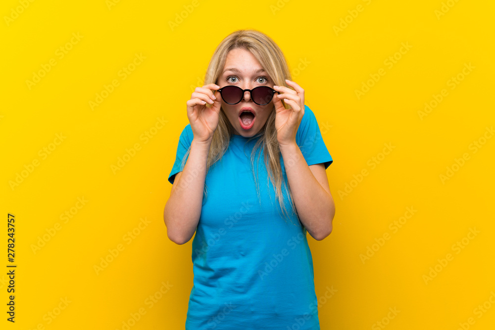 Young blonde woman over isolated yellow background with glasses and surprised