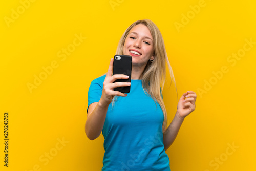 Young blonde woman over isolated yellow background making a selfie