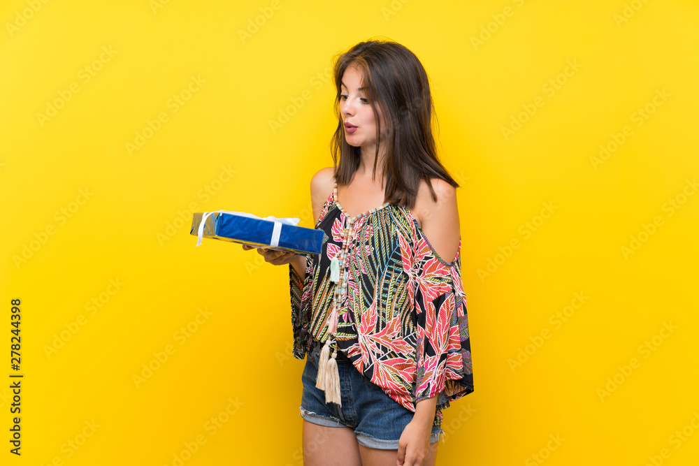 Caucasian girl in colorful dress over isolated yellow background holding gift box Caucasian girl in colorful dress over isolated yellow background holding gift box