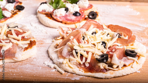 Cooking traditional Italian mini pizza, pizzetta - raw mini pizza with tomato sauce, black olives, jamon and mozzarella cheese on a cutting board, close-up