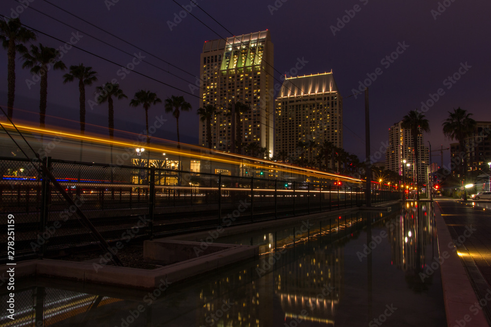 Streaks of Light from a Passing Trolley in the City of San Diego, California at Night - with City Lights Reflected in the Water, and Rows of Buildings and Palm Trees in the Background Streaks of Light from a Passing Trolley in the City of San Diego, California at Night - with City Lights Reflected in the Water, and Rows of Buildings and Palm Trees in the Background