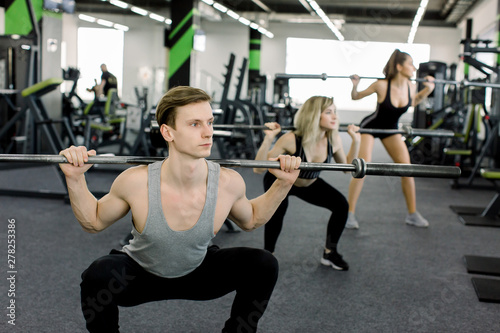 Young people working out with barbells at gym. Attractive women and handsome muscular man trainer are training in light modern gym. Beautiful girl squats under the supervision of the coach.