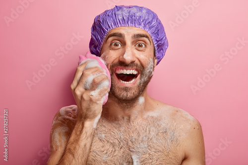 Naked glad man takes shower with soap sponge, washes face and torso, cares about body, wears bath cap on head, isolated on pink background. Washing, cleanliness, masculinity and hygiene concept