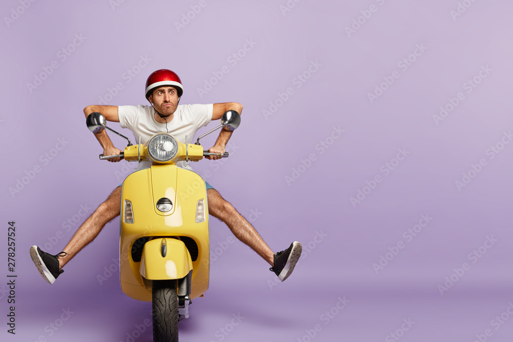Funny young man with fasten motorbike helmet, poses on fast bike, wears white t shirt and sneakers, poses against purple background with empty space. People, transportation and riding concept - obrazy, fototapety, plakaty 
