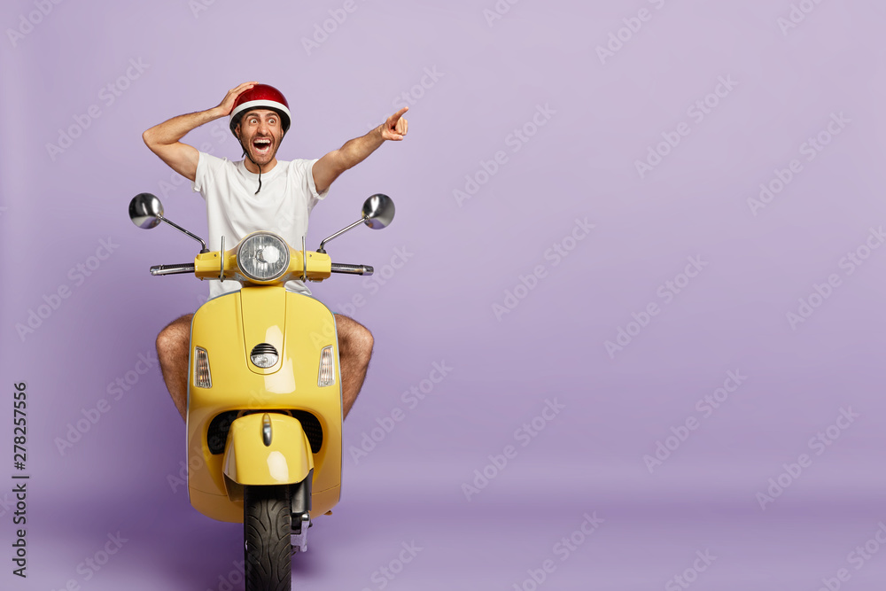 Surprised active man rides fast motorbike, points into distance, stares with shock, notices something incredible, wears protective helmet, isolated on purple background with blank copy space - obrazy, fototapety, plakaty 