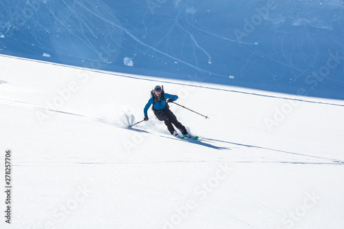 Backcountry skier on fresh powder slope in Alaska. Roller-balls fall behind him from steep cliff above.