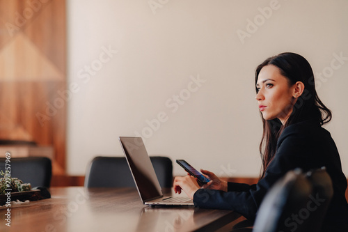 Young attractive emotional girl in business-style clothes sitting at a desk on a laptop and phone in the office or auditorium