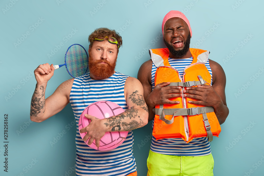 Sorrowful black man being in low spirit, poses in lifejacket, spends time  with redhaired friend who holds tennis racket and beach ball, play active  games on beach, stand against blue background Stock