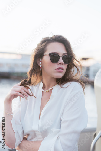 A beautiful girl in dark sunglasses and with beautiful styling is standing on the pier by the river. Girl in pearl jewelry and glasses