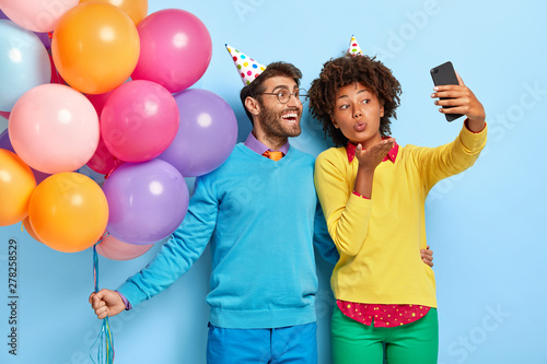 Smiling beautiful Afro girl and her happy boyfriend holds bunch of colorful air balloons, take selfie on smartphone, woman blows airkiss, wear party hats, enjoy festive occasion, pose indoor