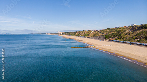 An aerial view of a majestic sandy beach with crystal blue water sea, groynes (breakwaters) and beach huts along a beautiful cliff with green vegetation under a blue and sunny sky
