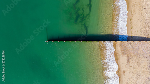 An aerial view of a groyne (breakwater) dividing a majestic cystal clear green water sea with crashing waves on a sunny sandy beach photo
