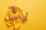 Handsome positive guy points away on right side, shows free space for your promotional information, stands in paper hole, wears yellow casual outfit and hat. People, advertisement, good mood concept