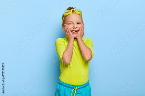 Charming lovely preschooler keeps hands on cheeks, enjoys swimming in pool, wears goggles on head, green casual t shirt and blue shorts, has fun during rest at summer. Playful girl learns to swim