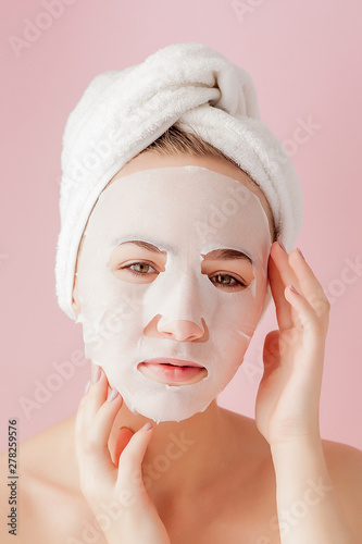 Beautiful young woman is applying a cosmetic tissue mask on a face on a pink background. Healthcare and beauty treatment and technology concept