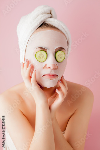 Beautiful young woman is applying a cosmetic tissue mask on a face with cucumber on a pink background