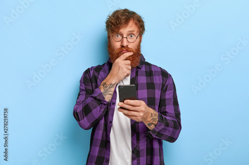 Photo of young redhead guy uses cellphone for online communication, looks with surprisement at camera, wears fashionable outfit and optical glasses, poses against blue background. Blogger with device photo