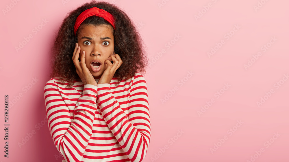 Stupefied panicking Afro woman poppes eyes, watches scary movie, keeps hands on cheeks, has frightened facial expression, dressed in casual outfit, isolated on pink wall with free space for text Stupefied panicking Afro woman poppes eyes, watches scary movie, keeps hands on cheeks, has frightened facial expression, dressed in casual outfit, isolated on pink wall with free space for text