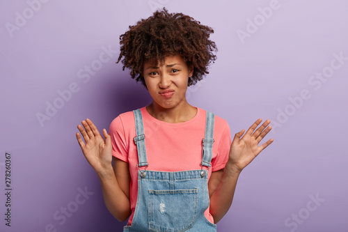 Thank yoy but no. Displeased dark skinned woman receivs disturbing suggestion, waves palms in rejection, grimaces unpleasantly, wears casual pink t shirt and dungarees, poses in studio alone