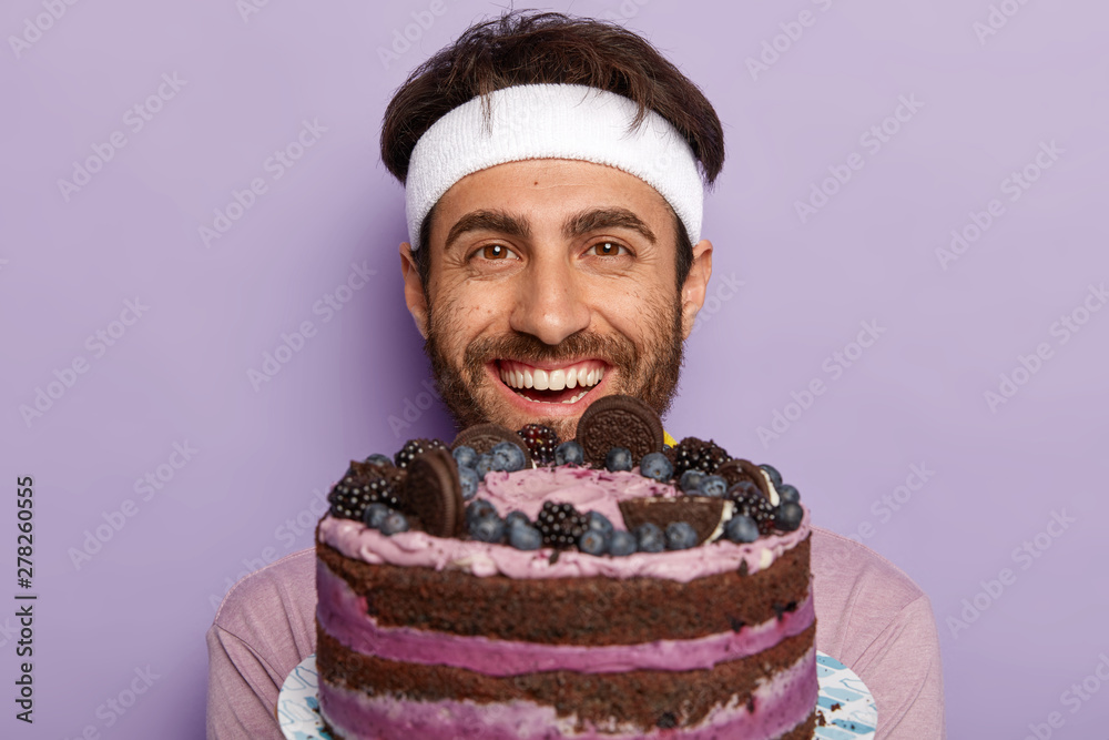 Headshot of handsome unshaven man glad to receive big delicious fruit cake, wants to eat something tasty after burning calories in gym, has toothy smile, poses indoor against violet studio wall. Headshot of handsome unshaven man glad to receive big delicious fruit cake, wants to eat something tasty after burning calories in gym, has toothy smile, poses indoor against violet studio wall.