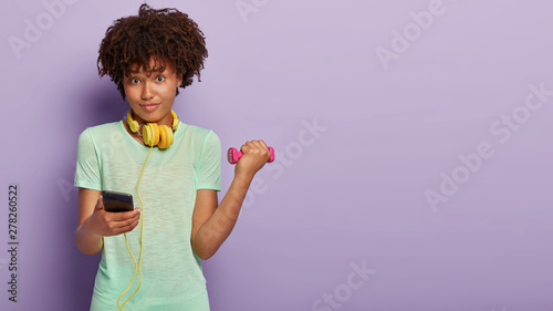 Technology and bodybuilding concept. Lovely healthy lady with curly hairstyle, holds dumbbell, works on muscles, uses smart phone for chatting online, listening music during training, likes fitness