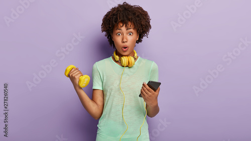 Sport, modern technologies and physical activity concept. Attractive Afro woman being speechless, keeps mouth opened, raises arm with dumbbell, strengthens biceps, shocked read news via cellular