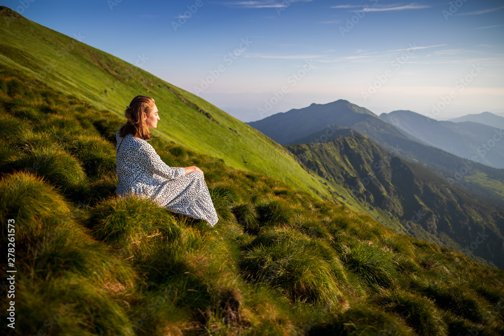 Naklejka premium Young woman sitting on a rock and looking to the horizon. Happy traveler enjoying sunset view. Girl in long white dress in the mountains