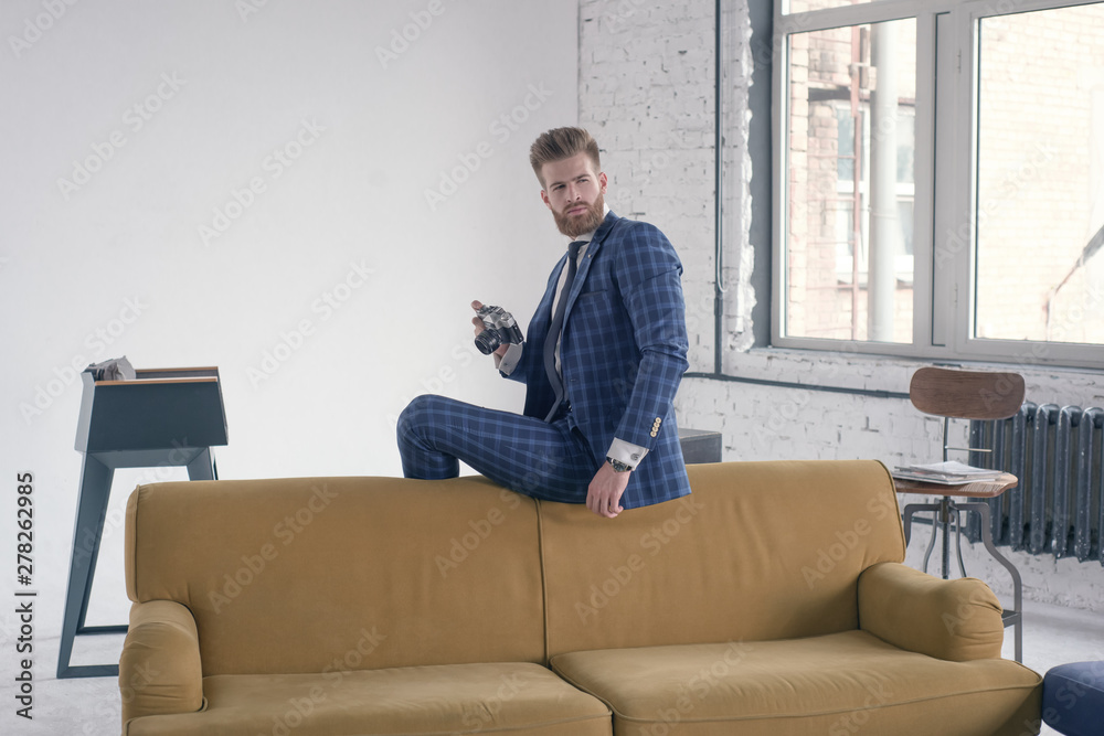 Handsome photographer. Good looking young man in full suit holding photographic camera and looking away while sitting on the sofa Handsome photographer. Good looking young man in full suit holding photographic camera and looking away while sitting on the sofa