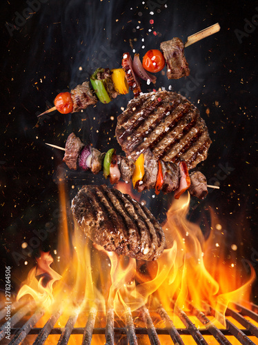Tasty beef steaks and skewers flying above cast iron grate with fire flames. Freeze motion barbecue concept.