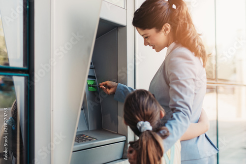 Fotografie Young mother elegantly dressed with her daughter using ATM machine on city street