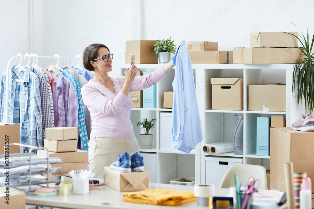 Office worker with smartphone taking picture of new stylish blue shirt on hangers for one of clients Office worker with smartphone taking picture of new stylish blue shirt on hangers for one of clients