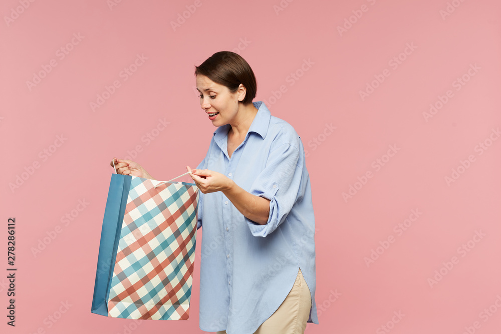 Young excited female in casualwear looking at surprise in open paperbag while posing in isolation Young excited female in casualwear looking at surprise in open paperbag while posing in isolation