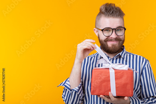 Surprised young hipster man with mustache and beard in surprise unpacks a red box with gifts on a yellow background with copy space, close up. The concept of gifts and surprises for the holiday © satura_