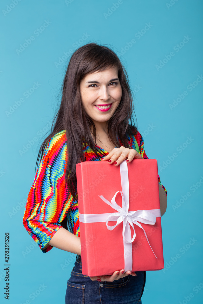 Cheerful good looking young brunette woman holding a red gift box in her hands, but against a blue background. Concept holidays and presents Cheerful good looking young brunette woman holding a red gift box in her hands, but against a blue background. Concept holidays and presents