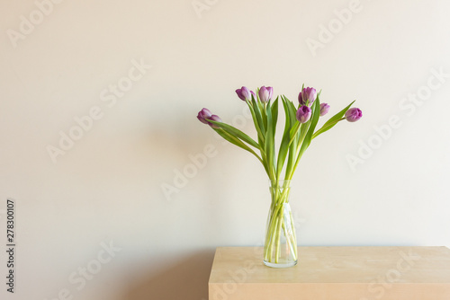 Closeup of purple tulips in glass vase on shelf against neutral wall background