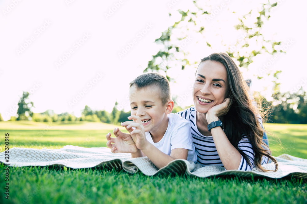 Happy family day. Beautiful lovely young mother with her cute little son is have a fun and hugging each other while lying on grass in park outdoors Happy family day. Beautiful lovely young mother with her cute little son is have a fun and hugging each other while lying on grass in park outdoors