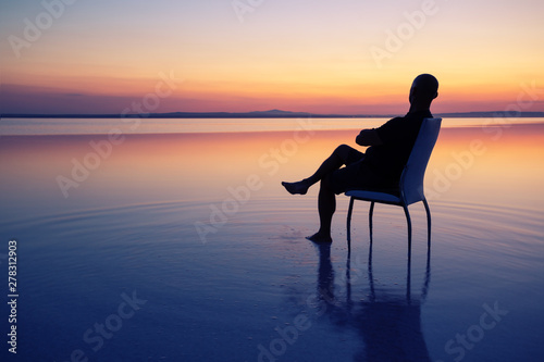 Silhouette of man sitting on chair in middle of lake at sunset