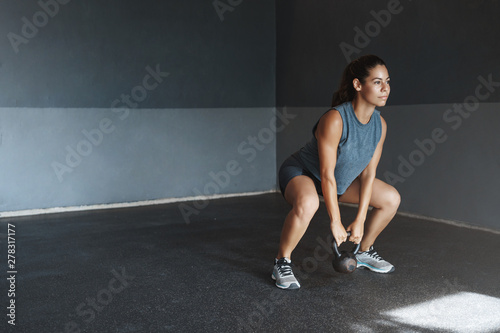 One more sit-up to achieve goal. Good-looking motivated hispanic sportswoman wear activewear, look determined hold kettlebell, make fitness crossfit exercise, perform squats stand gym