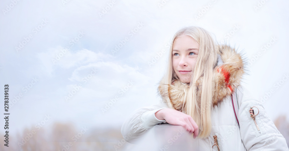 girl blonde in warm coat looks into distance standing on bridge with railing, sky background girl blonde in warm coat looks into distance standing on bridge with railing, sky background