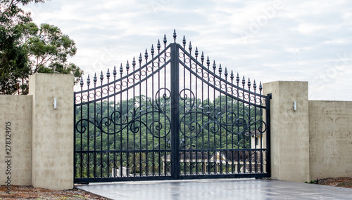 Black metal wrought iron driveway property entrance gates set in concrete brick fence, lights, garden trees in background