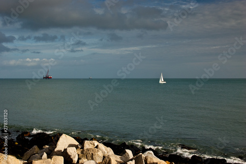 Small white sail yacht sailing in blue sea and oil platform, dramtic sky photo