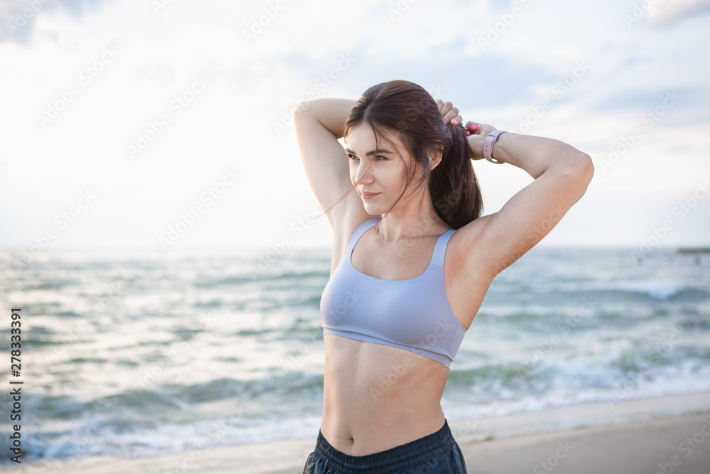 Pretty brunette woman fixes her hair after workout at the sea shore at sunrise. Model listening to the music during exercising near the sea. Pretty brunette woman fixes her hair after workout at the sea shore at sunrise. Model listening to the music during exercising near the sea.