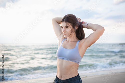 Pretty brunette woman fixes her hair after workout at the sea shore at sunrise. Model listening to the music during exercising near the sea.