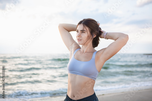 Pretty brunette woman fixes her hair after workout at the sea shore at sunrise. Model listening to the music during exercising near the sea.