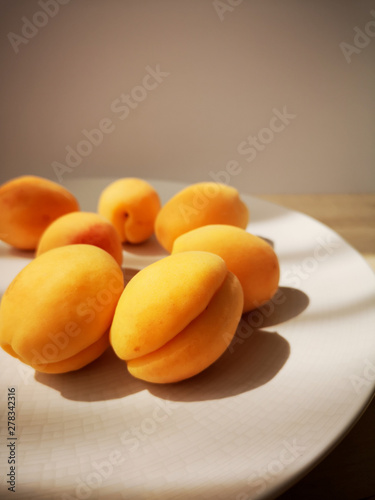 Several ripe juicy fragrant apricots lie on a large round white plate, which stands on a simple wooden table. Hard light.