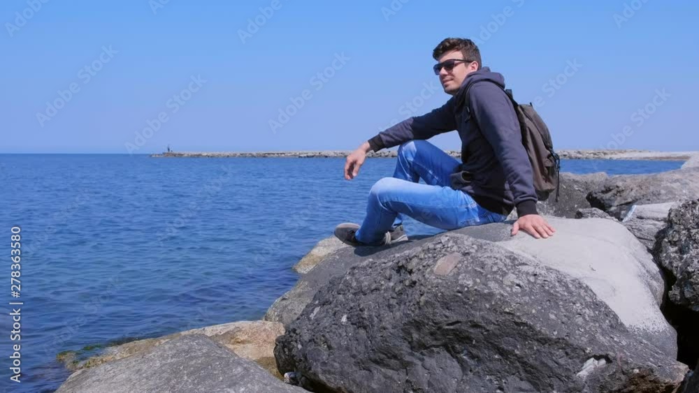 Young man tourist admires seascape sits at huge stone on the sea shore. Guy traveller in sunglasses, jeans and sweatshirt on sea spring vacation. Travel journey trip with backpack. Young man tourist admires seascape sits at huge stone on the sea shore. Guy traveller in sunglasses, jeans and sweatshirt on sea spring vacation. Travel journey trip with backpack.