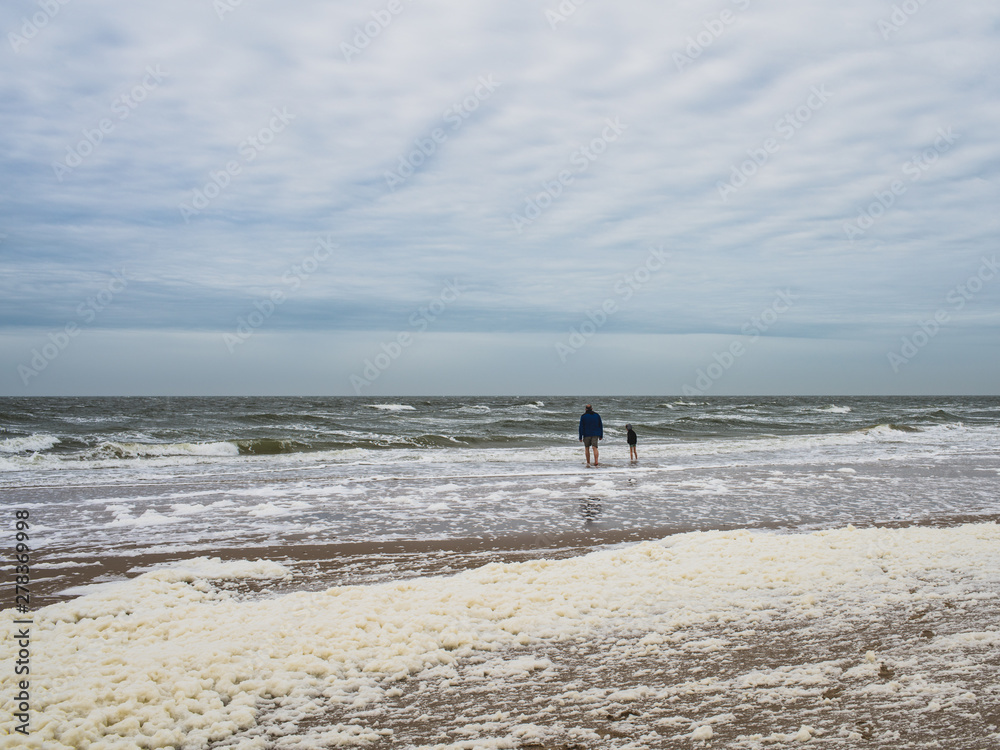 Grandfather and grandson stand on the beach and watch strong waves with foam. Egmond aan Zee, Netherlands, Europe