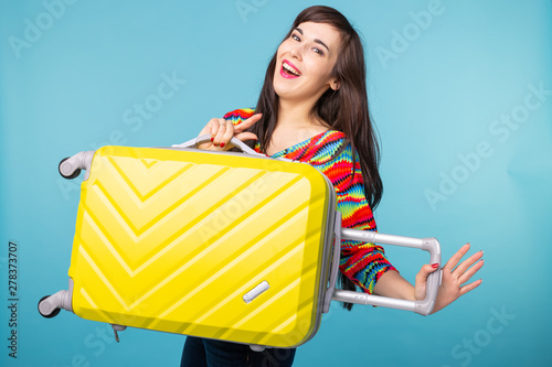 Joyful smiling young brunette woman posing with a yellow suitcase while waiting for a vacation. The concept of tourism and travel